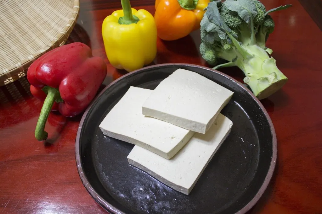Three slices of tofu on a round black plate with various colored bell peppers in the background.